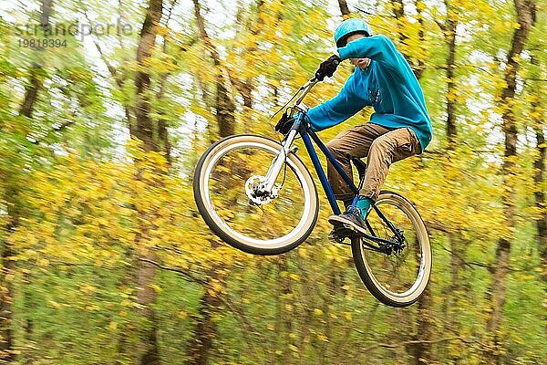 Ein junger Fahrer mit Helm und blauem Sweatshirt fliegt auf einem Fahrrad  nachdem er von einem hohen Kicker auf einem Waldradweg gesprungen ist. Aufnahme mit Langzeitbelichtung Verdrahtung