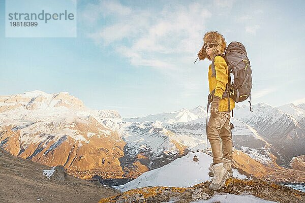 Das Backpacker Mädchen mit Sonnenbrille und einer großen nordischen Pelzmütze  das einen Rucksack auf dem Rücken trägt  steht auf einem Felsen und blickt in Richtung der Klippen  die sich im Felsen verstecken. Das Konzept der Erholung in den Bergen und des Tourismus. Urlaub in den Bergen. Ansicht von hinten