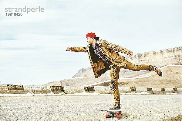 Stylish happy Junger Mann in Mütze und Hose Jogger rollen auf einem Longboard eine Bergstraße hinunter  genießen das Leben