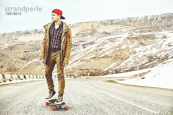 Stylish happy Junger Mann in Mütze und Hose Jogger rollen auf einem Longboard eine Bergstraße hinunter  genießen das Leben