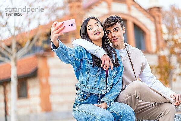 Multiethnisches Teenagerpaar macht ein Selfie im Sitzen und in Umarmung in einem Stadtpark