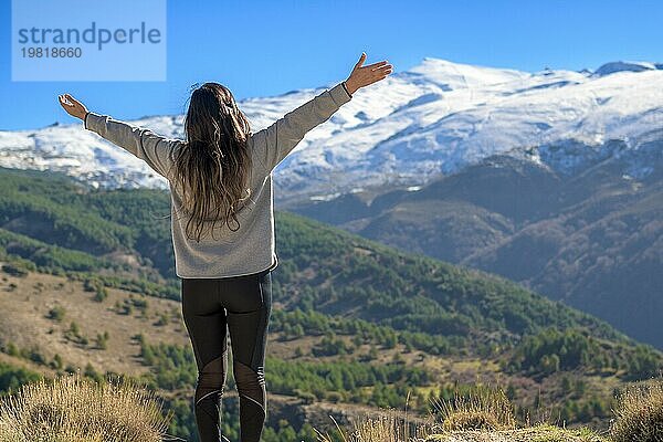 Eine Frau steht mit erhobenen Armen vor einer Berglandschaft mit einem schneebedeckten Gipfel unter einem klaren Himmel