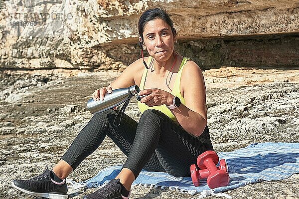 Lateinamerikanische Frau  mittleres Alter  ruht sich aus  stärkt sich  isst  trinkt Wasser  nach einem Fitnessstudio  verbrennt Kalorien  hält sich fit  draußen am Meer  trägt Kopfhörer und Smartwatch