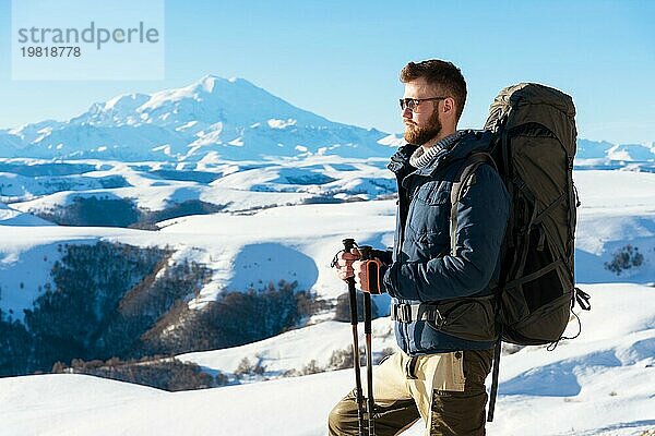 Ein Hipster Reisender mit Bart und Sonnenbrille in der Natur. Ein Mann wandern in den Bergen mit einem Rucksack und skandinavischen Wanderstöcke im Hintergrund eines Berges Elbrus Landschaft und blaün Himmel. Reisen Lifestyle Abenteuer Outdoor Erholung übertreffen