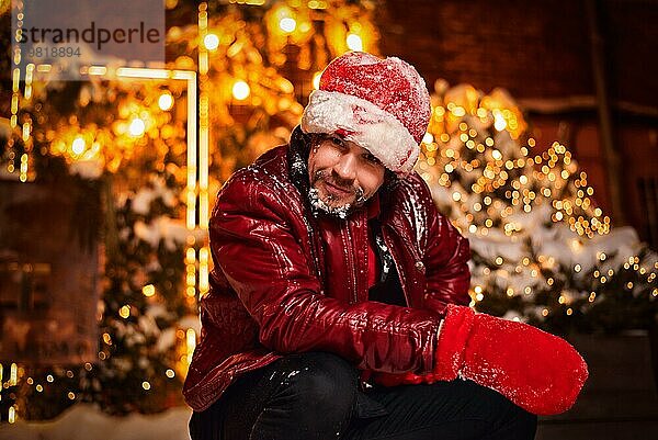 Mann gekleidet als Weihnachtsmann posiert auf einer Straße mit Girlanden geschmückt. Konzept für Neujahr und Weihnachten. Gemischte Medien