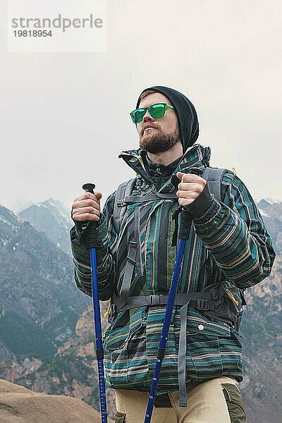 Ein Mann mit Bart und Sonnenbrille in einer Membranjacke  Hut  mit einem Rucksack und Stöcken für Nordic Walking  ein Reisender  der in der freien Natur steht und auf die Berge schaut