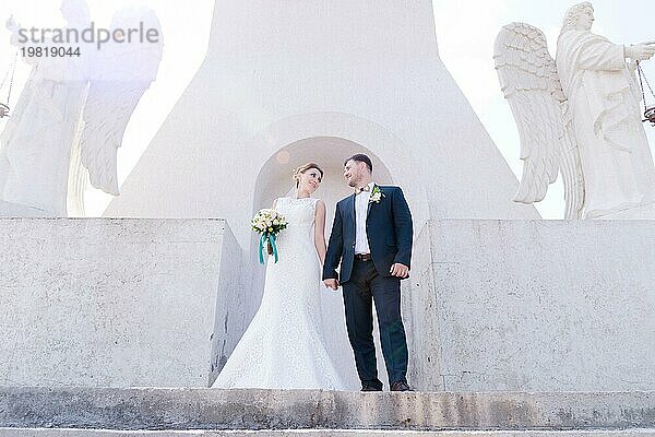 Porträt eines schönen Paares in den Flitterwochen an einem Hochzeitstag mit einem Blumenstrauß in der Hand vor dem Hintergrund eines orthodoxen christlichen Denkmal mit Engeln. Das Konzept der christlichen Hochzeit in der Kirche der Frischvermählten und der spirituelle Glaube der jungen Familie