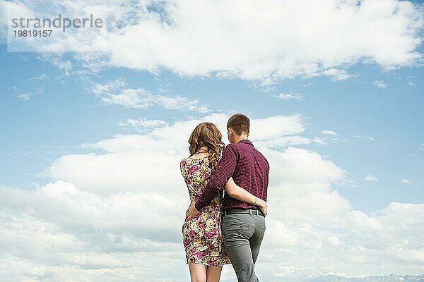 Blick von hinten auf ein junges Paar  das in einer Umarmung steht und in die Ferne gegen den Wolkenhimmel blickt. Eine Vision für die Zukunft