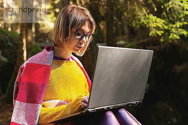 Ein getöntes Porträt eines lächelnden Freelancer Hipster Mädchens mit Brille  das in eine Decke gekleidet ist und mit einem Laptop auf einem Felsen in einem Nadelwald im warmen Sonnenlicht kniet. Das Konzept der freiberuflichen Arbeit in einem Abstand und Reisen ohne Unterbrechung w