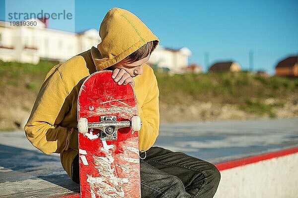 Nahaufnahme eines jungen Hipsters in Sweatshirt und Jeans  der traurig in einem Skatepark sitzt und ein Skateboard hält