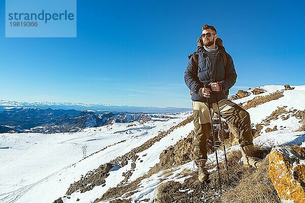 Ein Hipster Reisender mit Bart und Sonnenbrille in der Natur. Ein Mann wandert in den Bergen mit einem Rucksack und skandinavischen Wanderstöcken vor dem Hintergrund einer Berglandschaft und blauem Himmel. Reisen Lifestyle Abenteuer Outdoor Erholung Outdoor Sport