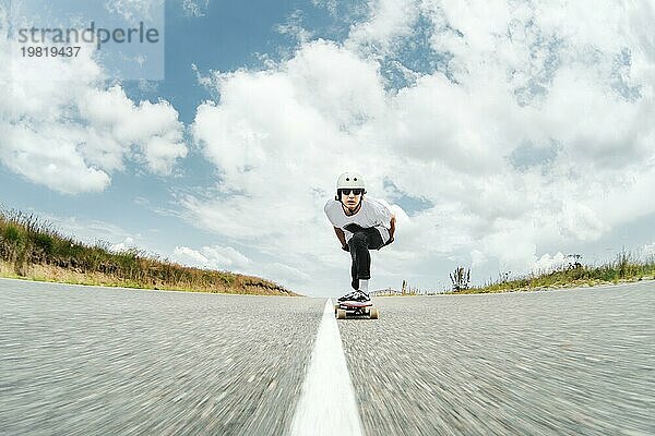 Der Typ mit dem Helm und der Sonnenbrille fährt mit seinem Longboard in einem speziellen Sportständer sehr schnell über die asphaltierte Straße