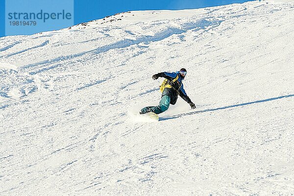 Ein Freeboard Snowboarder mit Skimaske und Rucksack fährt über die schneebedeckte Piste und hinterlässt einen Pulverschnee vor dem blaün Himmel