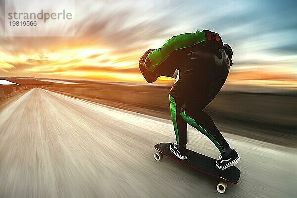 Ein Mann in Helm und Lederkombi fährt in einem speziellen Gestell mit hoher Geschwindigkeit auf einem Longboard bergab auf Afsalt vor der Kulisse eines schönen Sonnenuntergangshimmels im Gegenlicht der untergehenden Sonne. Der Hintergrund ist dynamisch aufgefächert