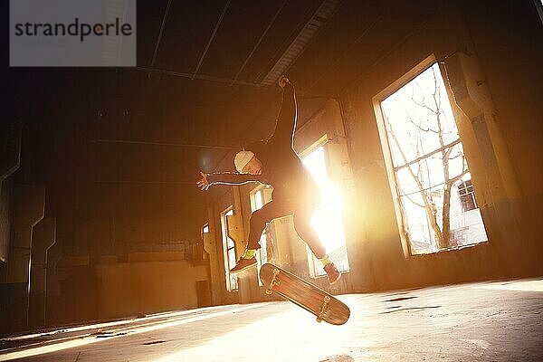 Ein junger Skater mit weißer Mütze und schwarzem Sweatshirt macht einen Trick mit einem Skatesprung in einem verlassenen Gebäude im Gegenlicht der untergehenden Sonne. Das Konzept der jugendlichen Untergrundkultur und des Skateboarding