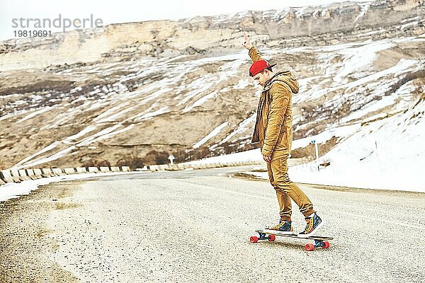 Stylish happy Junger Mann in Mütze und Hose Jogger rollen auf einem Longboard eine Bergstraße hinunter  genießen das Leben