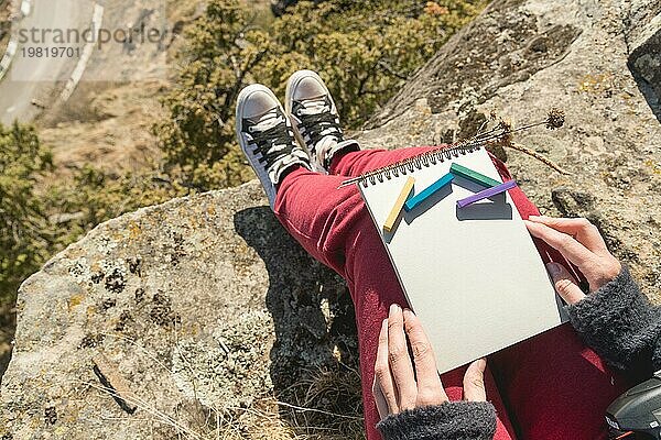 Blick aus der ersten Person auf den weiblichen Schoß ist ein Notizbuch mit Kreiden für die Zeichnung Pastelle. Weibliche Hände halten ein Notizbuch in der Natur. Ein Mädchen  das auf einer Klippe in der Nähe einer Klippe sitzt  malt eine Pastelllandschaft. Beschaffung für den Designer