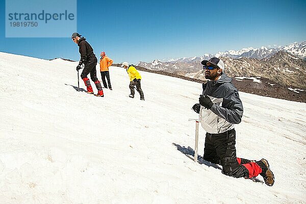 Training zum Korrigieren des Ausrutschens an einem Hang oder auf einem Gletscher mit Hilfe eines Eispickels. Ein voll ausgerüsteter Rucksacktourist kniet auf einem verschneiten Hang in den Bergen in der Nähe des Eispickels. Im Hintergrund beobachten Bergsteiger  die einen unerfahrenen Kletterer vor dem Aufstieg trainieren