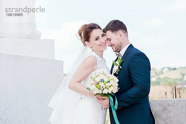 Porträt eines schönen Paares in den Flitterwochen an einem Hochzeitstag mit einem Blumenstrauß in der Hand vor dem Hintergrund eines orthodoxen christlichen Denkmal mit Engeln. Das Konzept der christlichen Hochzeit in der Kirche der Frischvermählten und der spirituelle Glaube der jungen Familie