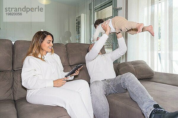 Die Mutter benutzt ein Tablet  während der Vater mit dem Baby spielt  das zusammen auf dem Sofa zu Hause sitzt