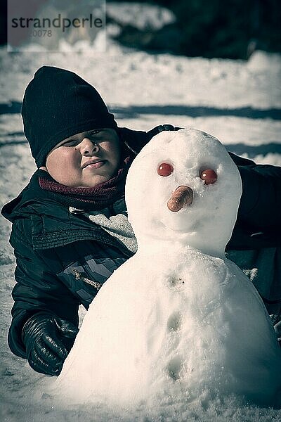 Junge liegt im Schnee neben einem selbstgebauten Schneemann mit Karottennase und Tomatenaugen