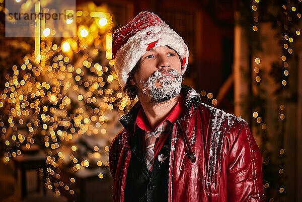 Ein als Weihnachtsmann verkleideter Mann posiert auf einer mit Girlanden geschmückten Straße. Sein Bart ist mit Schnee bedeckt. Konzept für Neujahr und Weihnachten. Gemischte Medien