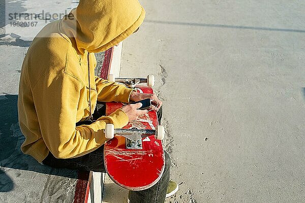 Nahaufnahme eines Teenagers in Sweatshirt  Jeans und Turnschuhen  der in einem Skatepark sitzt und ein Telefon mit Kopfhörern und ein Skateboard hält