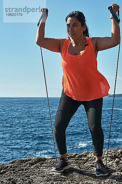 Lateinamerikanische Frau  mittleres Alter  in Sportkleidung  Training  körperliche Übungen  Muskulatur  mit Gummiband  Kalorien verbrennen  sich fit halten  draußen am Meer  mit Kopfhörer  Smartwatch