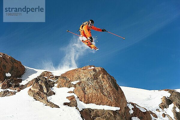 Ein professioneller Skifahrer springt von einer hohen Klippe gegen den blaün Himmel und hinterlässt eine Spur aus Pulverschnee in den Bergen. Foto von den Hängen des Berges Elbrus. Das Konzept des Extremsports und der Erholung in den Bergen im Winter. Kopieren Sie den Raum