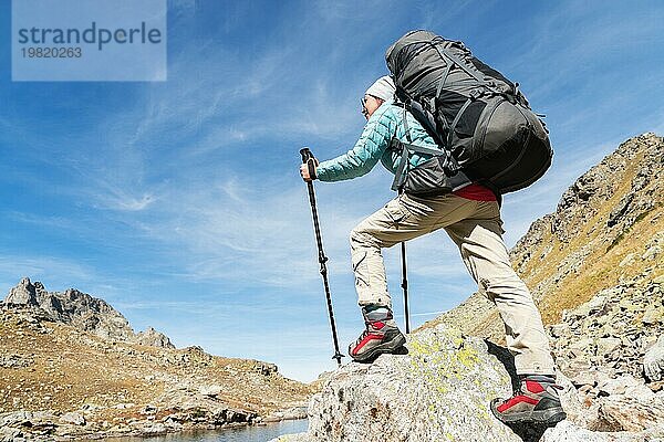 Schlankes und sympathisches Wandermädchen mit Sonnenbrille und blaür dünner Daunenjacke mit Rucksack und Trekkingstöcken steigt auf einen hohen Felsen vor dem Hintergrund von Felsen und einem hohen Bergsee