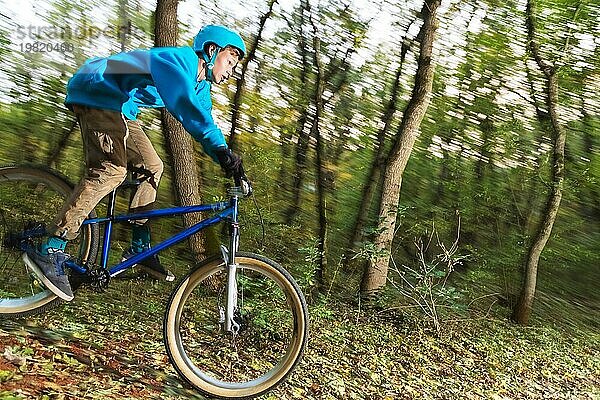 Ein junger Fahrer mit Helm und blauem Sweatshirt fliegt auf einem Fahrrad  nachdem er von einem hohen Kicker auf einem Waldradweg gesprungen ist. Aufnahme mit Langzeitbelichtung Verdrahtung