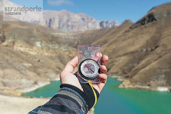 Ein Mann hält einen Taschenmagnetkompass zur Navigation vor dem Hintergrund eines felsigen Abhangs und eines Bergsees in der Hand. Das Konzept  einen Weg zu finden. Freiheit gewinnen. Freiheit der Wahl