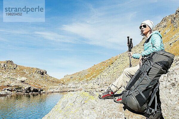 Ein Touristenmädchen mit Sonnenbrille  Daunenjacke und Hut  mit einem Rucksack und einer Bergausrüstung mit Griffen zum Wandern in den Händen  sitzt und entspannt sich und genießt die schöne Aussicht auf einen Hochgebirgssee im Nordkaukasus. Karatschai Tscherkess R