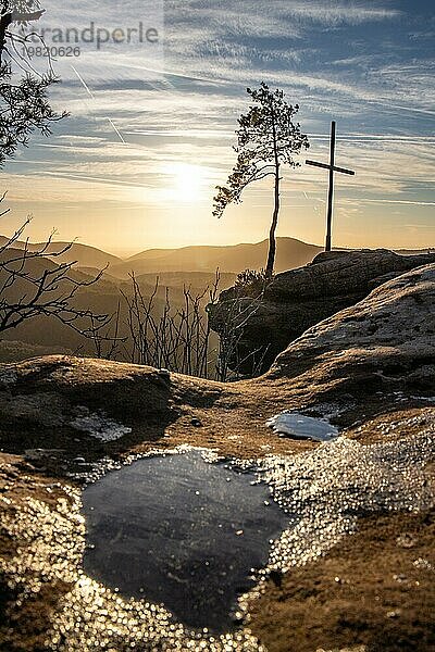 Landschaftsaufnahme auf einem Sandsteinfelsen im Wald. Morgenstimmung bei Sonnenaufgang an einem Aussichtspunkt. Ein kleiner Baum und ein Gipfelkreuz stehen auf dem Rötzenfels im Pfälzerwald