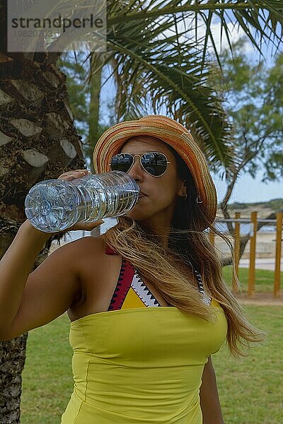 Glückliche Frau trinkt Mineralwasser neben einer Palme und genießt den Sommer. Kopierraum