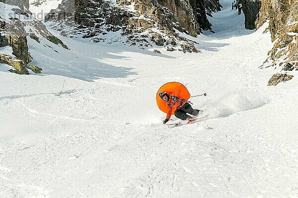 Ein männlicher Freerider mit Bart fährt im Backcountry mit hoher Geschwindigkeit von der Piste ab und hinterlässt eine Spur aus Pulverschnee mit dem geöffneten Lawinenkissen ABS vor dem Hintergrund der epischen Felsen. Das Konzept der Freeride Kultur und des Backcountry