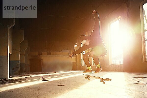 Ein junger Skater mit weißer Mütze und schwarzem Sweatshirt macht einen Trick mit einem Skatesprung in einem verlassenen Gebäude im Gegenlicht der untergehenden Sonne. Das Konzept der jugendlichen Untergrundkultur und des Skateboarding