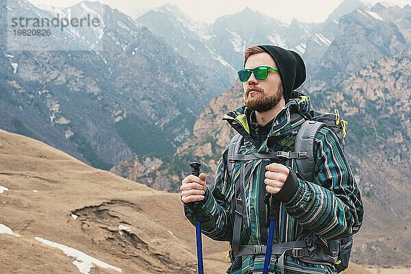 Ein Mann mit Bart und Sonnenbrille in einer Membranjacke  Hut  mit einem Rucksack und Stöcken für Nordic Walking  ein Reisender  der in der freien Natur steht und auf die Berge schaut