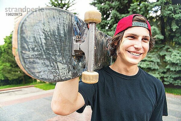 Porträt eines jungen  langhaarigen Skaters mit einer Mütze und seinem Lieblings Skateboard auf der Schulter