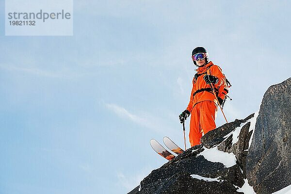 Ein professioneller Skisportler steht an einem sonnigen Tag am Rande eines hohen Felsens vor dem blaün Himmel. Kopieren Sie den Raum. Das Konzept des Skifahrens