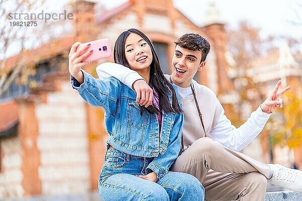 Fröhliches multiethnisches Paar  das auf der Straße sitzend ein Selfie mit dem Mobiltelefon macht