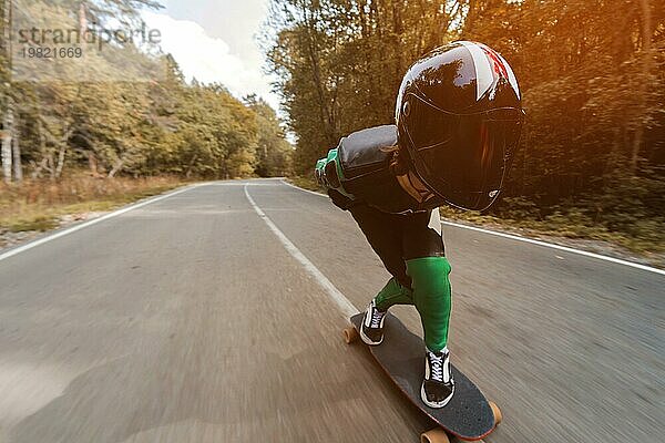 Ein junger Fahrer in einem speziellen Lederanzug und einem Ganzkörperhelm fährt mit hoher Geschwindigkeit auf seinem Longboard auf einer Landstraße im Herbstwald