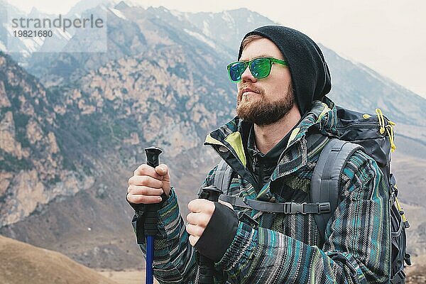 Ein Mann mit Bart und Sonnenbrille in einer Membranjacke  Hut  mit einem Rucksack und Stöcken für Nordic Walking  ein Reisender  der in der freien Natur steht und auf die Berge schaut