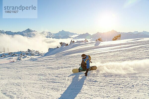 Ein Snowboarder mit Skimaske und Rucksack fährt auf einer schneebedeckten Piste und hinterlässt einen Pulverschnee vor dem blaün Himmel und der untergehenden Sonne. Foto in Bewegung