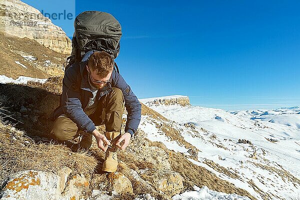 Porträt eines Hipster Reisenden mit Bart und Sonnenbrille sitzt und bindet Schnürsenkel in der Natur. Ein Mann wandert in den Bergen mit einem Rucksack und skandinavischen Wanderstöcken im Hintergrund einer Berglandschaft. Reisen Lifestyle Abenteuer ou