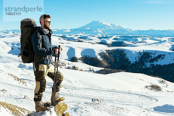 Ein Hipster Reisender mit Bart und Sonnenbrille in der Natur. Ein Mann wandern in den Bergen mit einem Rucksack und skandinavischen Wanderstöcke im Hintergrund eines Berges Elbrus Landschaft und blaün Himmel. Reisen Lifestyle Abenteuer Outdoor Erholung übertreffen