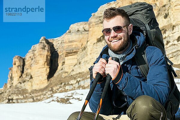 Porträt eines fröhlich lachenden Hipster Reisenden mit Bart und Sonnenbrille  der in der Natur sitzt. Ein Mann wandert in den Bergen mit einem Rucksack und skandinavischen Wanderstöcken im Hintergrund einer Berglandschaft. Reisen Lifestyle Abenteuer im Freien erholen