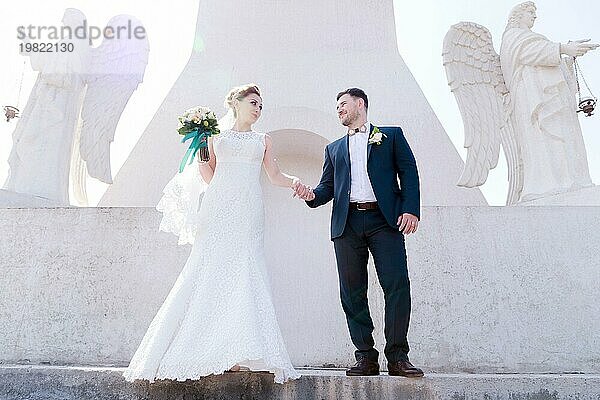 Porträt eines schönen Paares in den Flitterwochen an einem Hochzeitstag mit einem Blumenstrauß in der Hand vor dem Hintergrund eines orthodoxen christlichen Denkmal mit Engeln. Das Konzept der christlichen Hochzeit in der Kirche der Frischvermählten und der spirituelle Glaube der jungen Familie