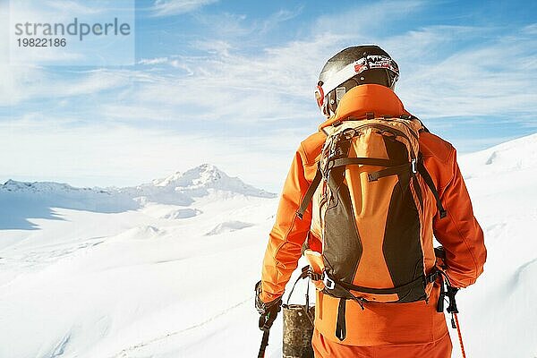 Ein Skifahrer in einem orangefarbenen Overall  mit einem Rucksack auf dem Rücken  einem Helm und Skistöcken in den Händen steht an einem Abgrund inmitten einer wunderschönen kaukasischen Berglandschaft mit schneebedeckten Gipfeln der Berge des Ca