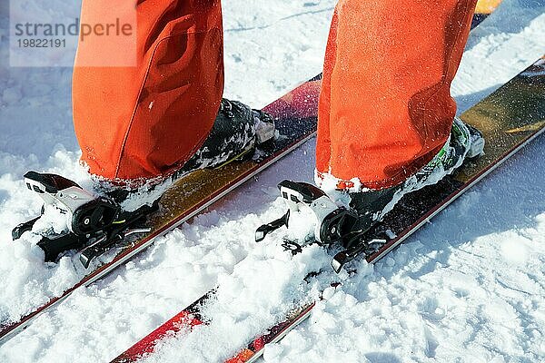 Orangefarbene Alpinskischuhe in einer Skihalterung. Ein Schuh ist vollständig auf den Skiern befestigt  der zweite nicht. Nahaufnahme. Orangefarbener Anzug